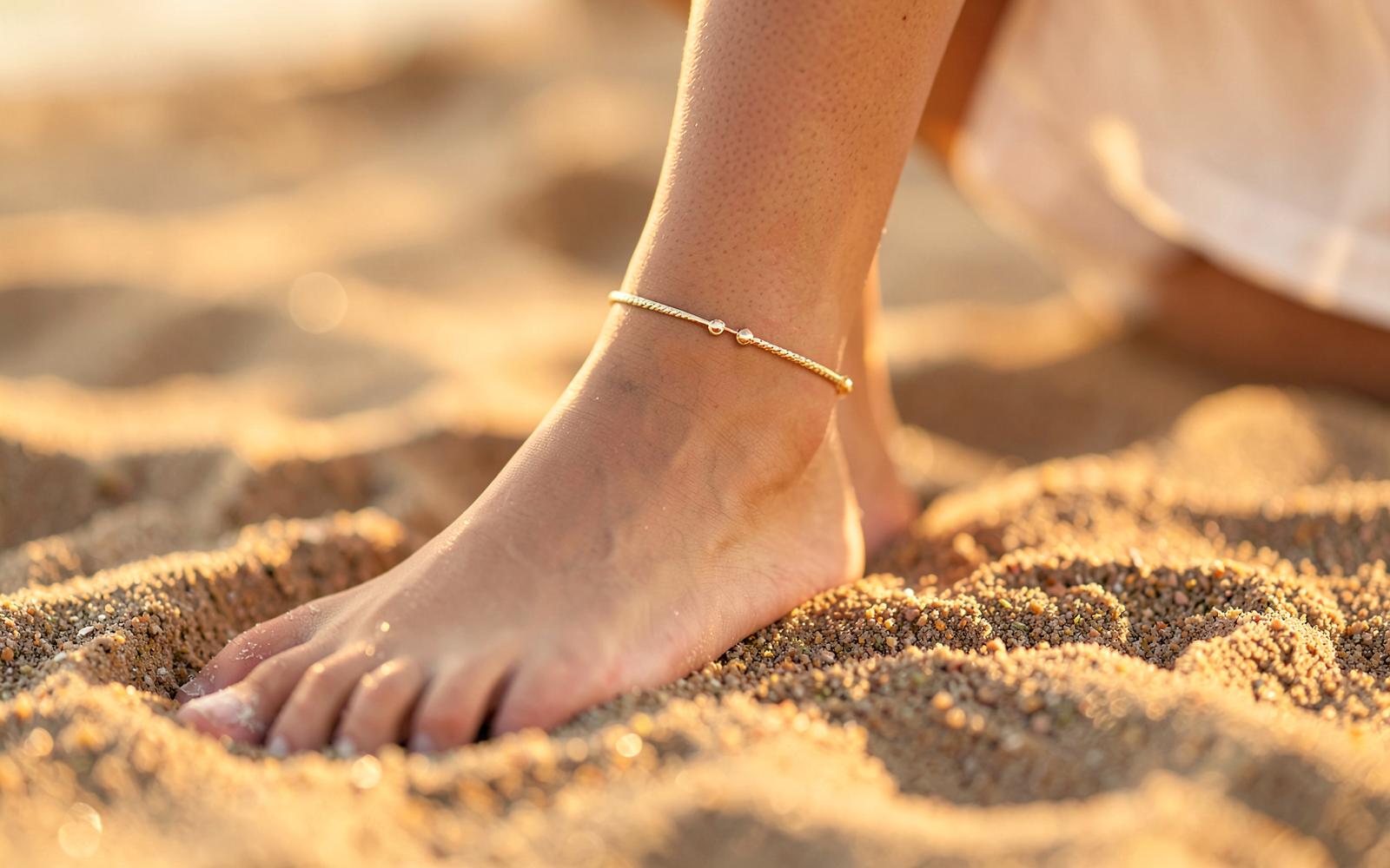 A delicate gold-tone anklet worn barefoot on warm sand at sunset